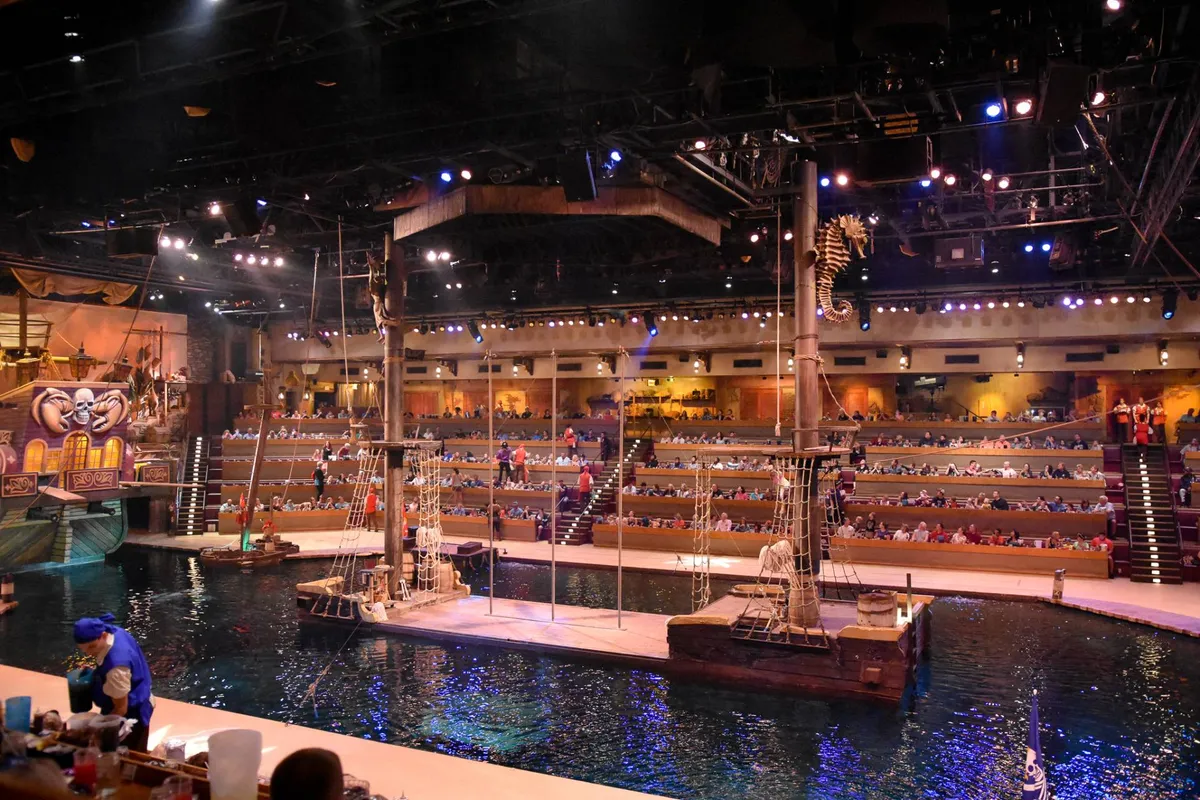 The image shows a large indoor aquatic stage set up for a live performance, featuring a ship structure surrounded by water, themed props, and rows of audience seating. The immersive atmosphere suggests entertainment and group experiences beyond the property itself.