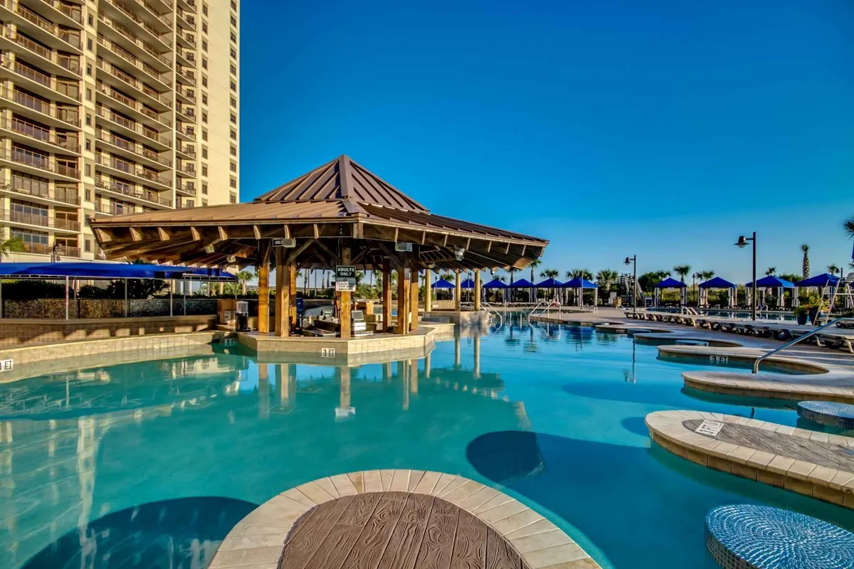 A lively resort pool area with multiple pools, palms, a sunlit pavilion, and plenty of lounge chairs. People are swimming, socializing, and enjoying drinks at the swim-up bar. The environment is vibrant, with activity for all ages and plenty of spaces to relax or play.