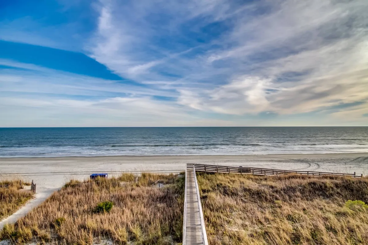A wide view of North Myrtle Beach with golden sands stretching towards the horizon, bordered by rolling dune grass. The vast blue ocean meets the clear sky, while a wooden boardwalk offers easy beach access. The scene is peaceful, ideal for barefoot walks, sandcastle building, and family fun by the shore.