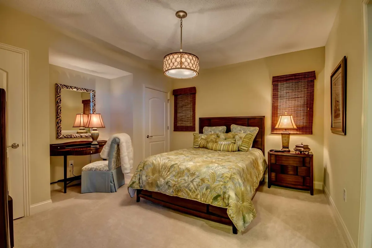 A guest bedroom highlighted by a bed with patterned bedding, a decorative lamp on the nightstand, and a mirror above the desk. The room is bathed in soft light, creating a cozy and calm ambiance ideal for relaxation or reading.