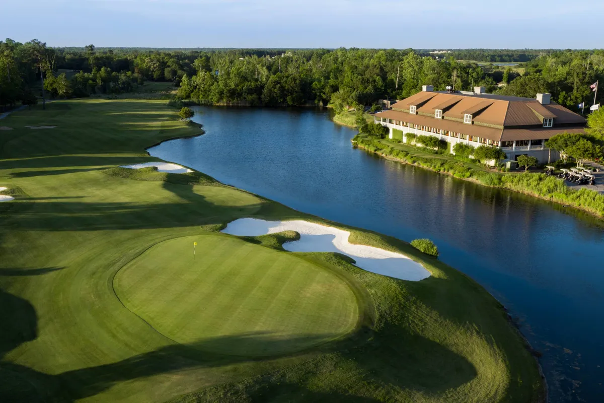 A wide, sunlit golf course stretches out with a gleaming lake beside it and a stately clubhouse in the distance. The green fairways are perfectly manicured, creating an inviting atmosphere for golfers and outdoor enthusiasts alike.