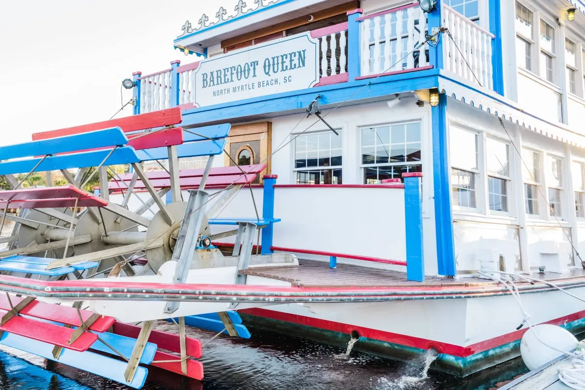 A charming paddlewheel riverboat is docked by the water, its colorful facade and prominent signage inviting guests aboard. The deck is set up for relaxed dining and social gatherings, capturing the festive spirit of a river cruise.