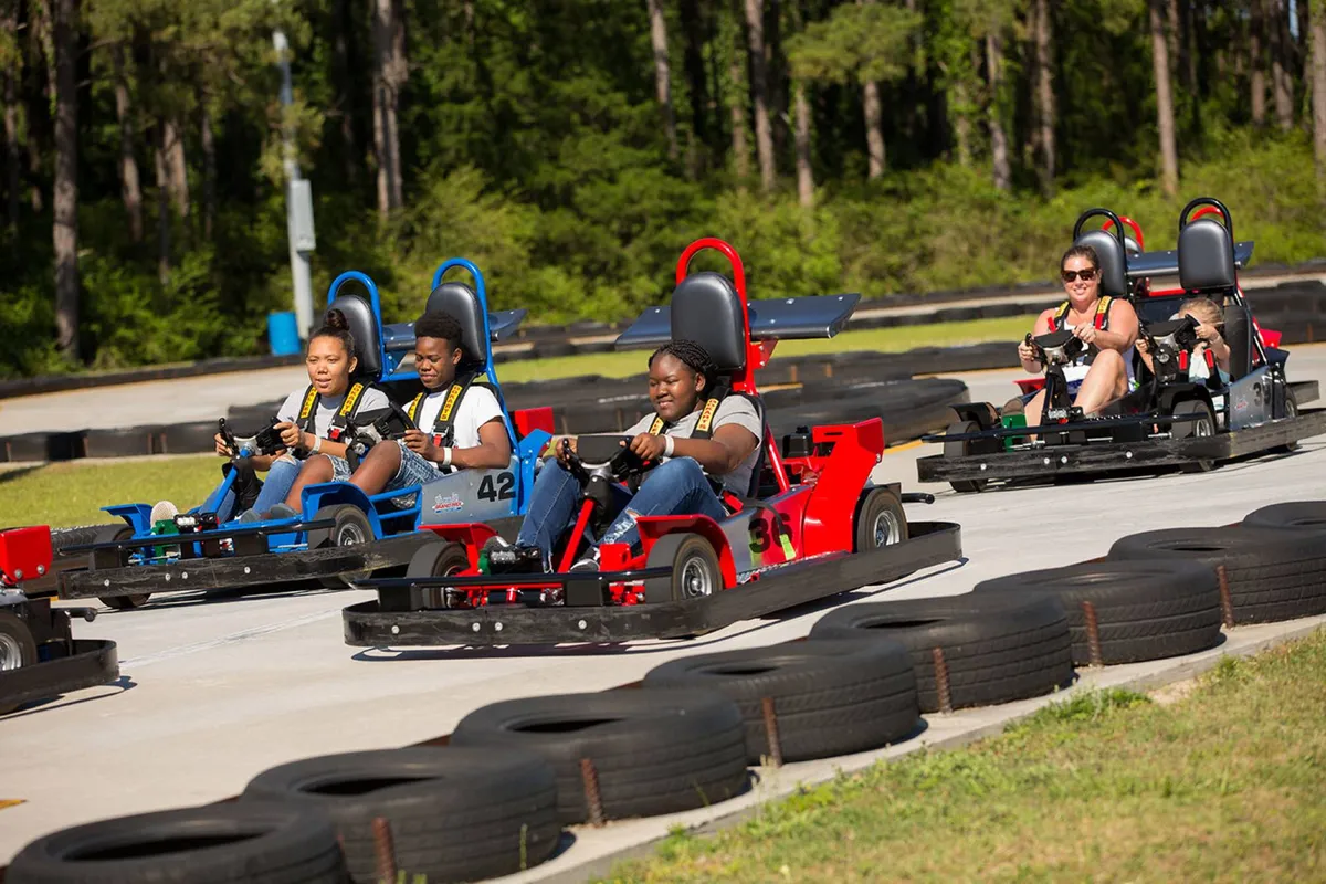 An action-filled go-kart race is underway on a vibrant outdoor track, with several participants zipping around a curve. Spectators watch excitedly, and the atmosphere is lively and energetic, perfect for families and thrill-seekers.
