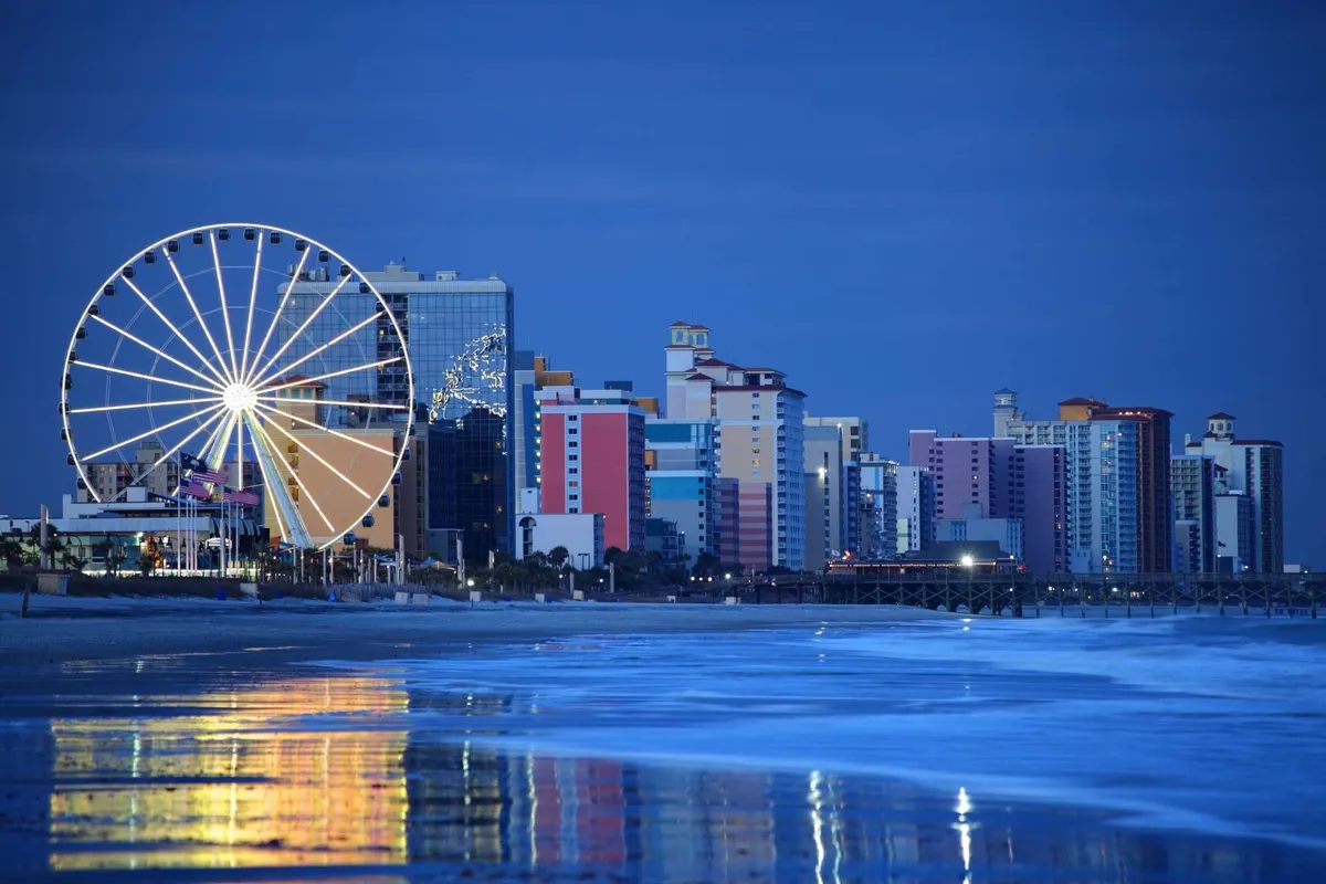 A festive coastal area with a large Ferris wheel, colorful waterfront buildings, and twinkling reflections on the water. The scene buzzes with activity, suggesting nearby attractions and evening entertainment options for all ages.