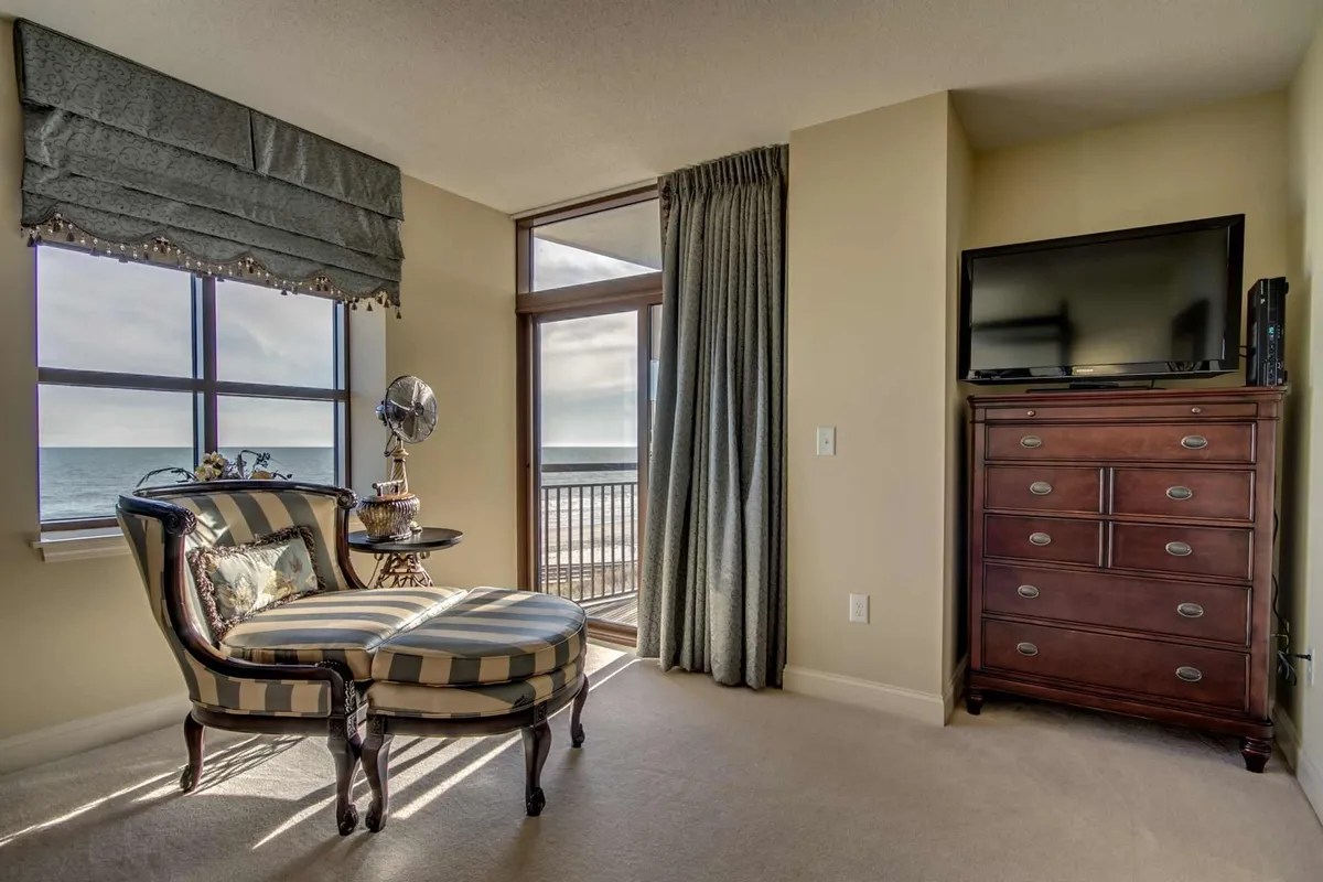 A cozy corner with a striped chair positioned beside a window overlooking the ocean. The scene includes a wooden dresser and TV, blending relaxation and entertainment with the ever-present serene water view.