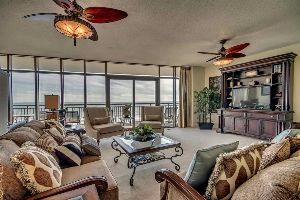 A spacious living room with large floor-to-ceiling windows that frame expansive ocean views. Comfortable sofas are arranged for both relaxation and conversation, while an elegant coffee table and a decorative ceiling fan add sophistication. The sunlight streaming in creates a serene and cozy atmosphere, perfect for group gatherings.
