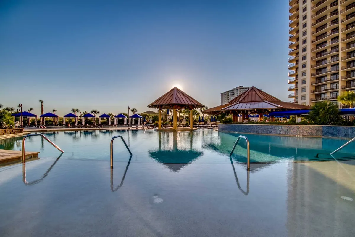 An inviting exterior shot featuring a luxurious swimming pool, pavilions with thatched roofs, numerous lounge chairs, and lush palm trees. The pool area enjoys tranquil surroundings with a high-rise resort building in the background, suggesting both exclusivity and convenience.