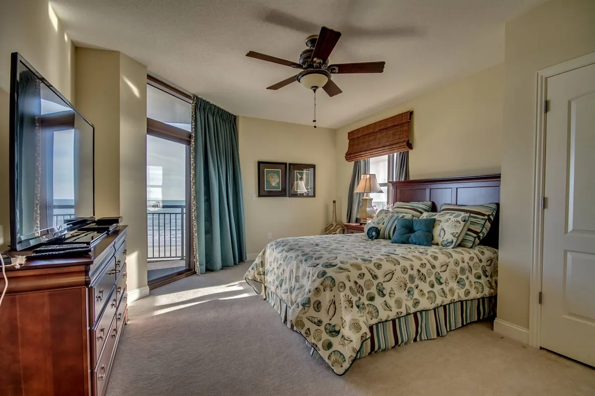 A beautifully styled bedroom featuring a large bed with decorative bedding, a spacious window framing a sea view, a ceiling fan, wooden dresser, and tasteful artwork on the walls. This room exudes tranquility and offers a glimpse of the ocean, suggesting a serene retreat.