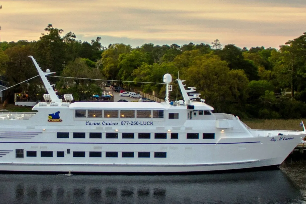 An exterior view of a luxurious cruise ship set against calm waters and lush greenery, with the sky painted in sunset hues. The scene exudes excitement and adventure, inviting guests to enjoy lively entertainment, dining, and nightlife.